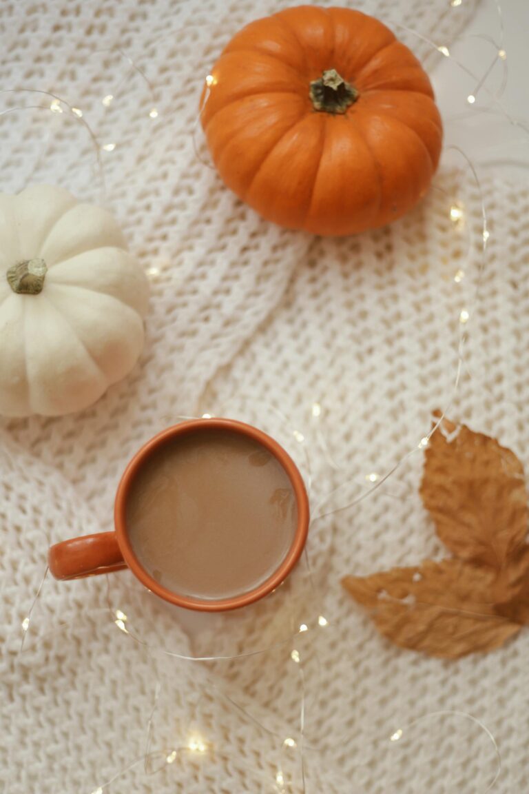 Warm autumn composition featuring coffee, pumpkins, and fairy lights on a knitted blanket.